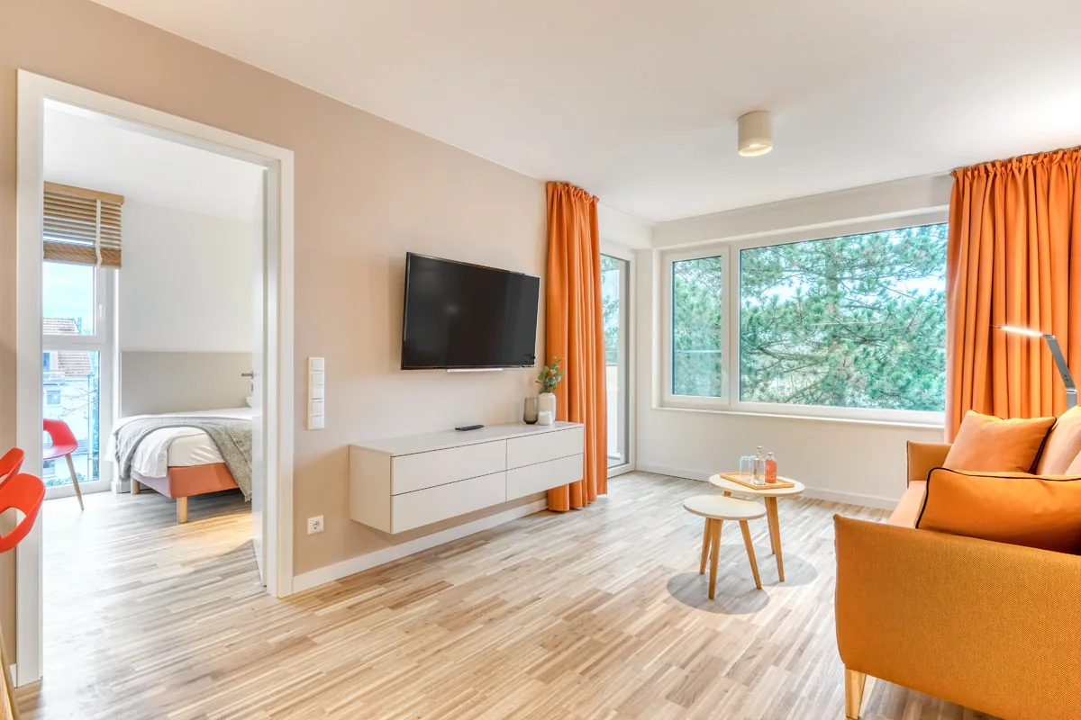 Wohnbereich eines Apartments mit orangefarbenen Vorhängen und Durchblick ins Schlafzimmer
