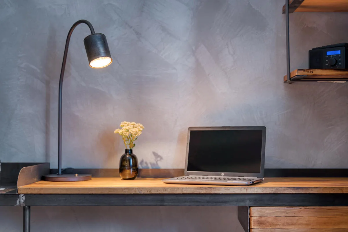 Arbeitsplatz vor Betonwand mit Leselampe, Laptop und Trockenblumen auf massiver Holzplatte