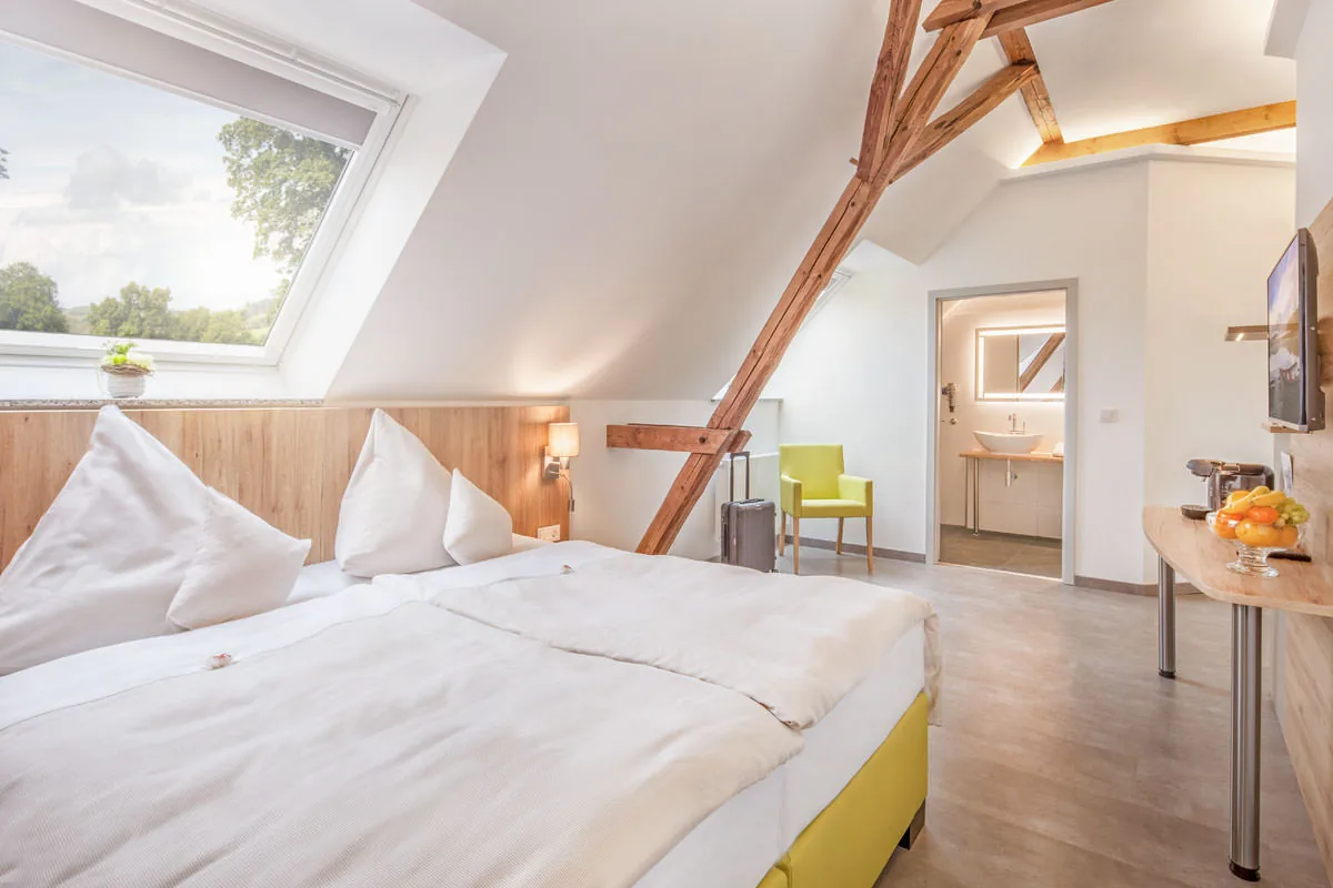 Dachzimmer mit freigelegten Holzbalken, Dachfenster und offenem Bad, gelber Sessel