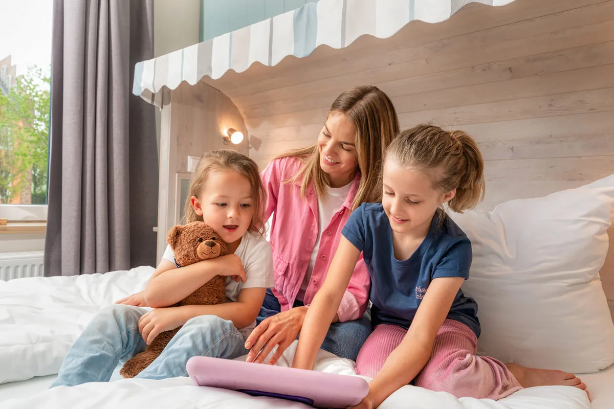 Mutter mit zwei Kindern auf dem Hausbett, Teddy und Tablet auf der Decke