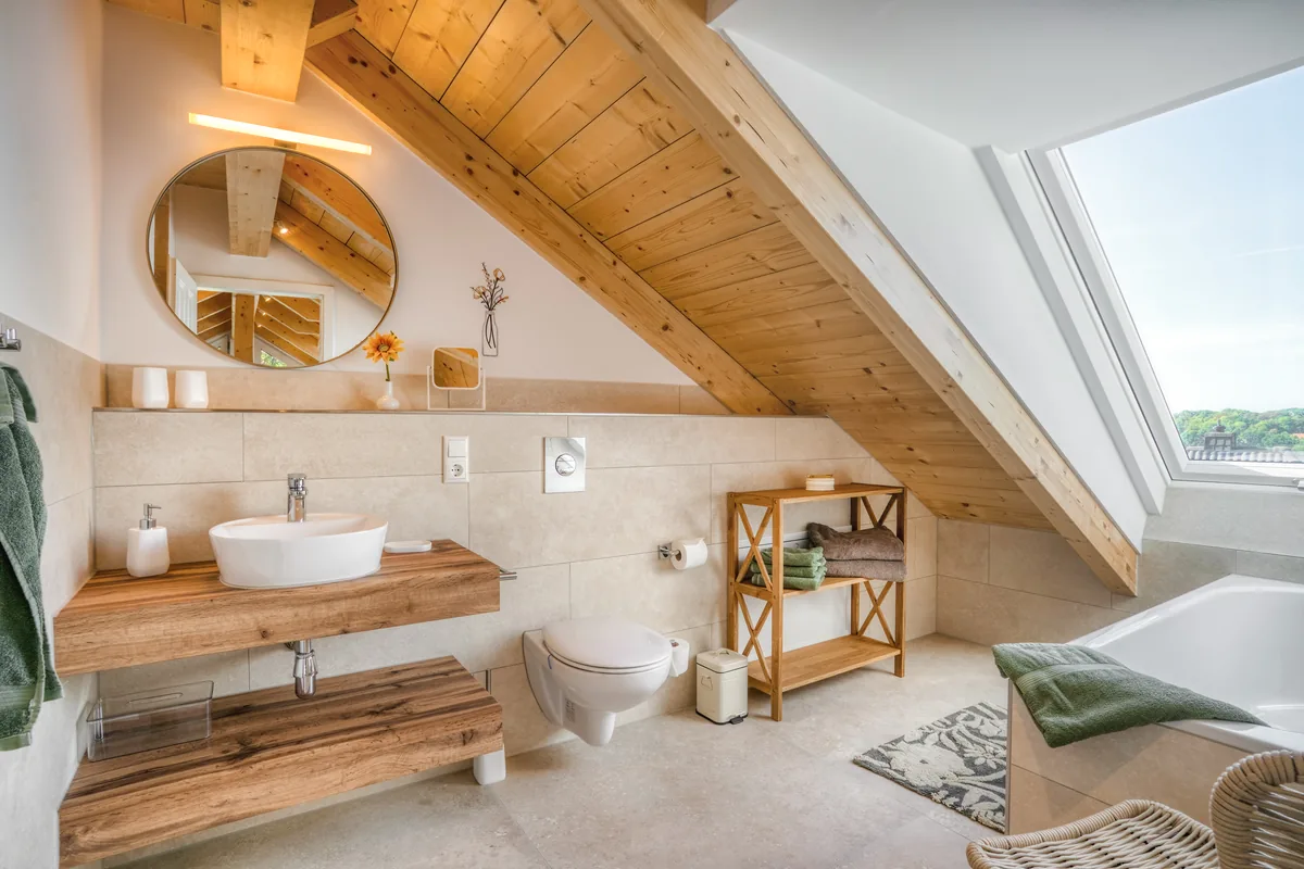 Bad im Ferienhaus unter der Dachschräge mit Rundspiegel, Holzwaschtisch und Wanne am Fenster