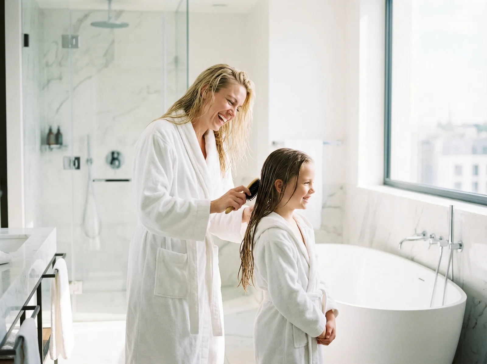 Mother combing her daughter's hair in a marble bathroom with freestanding tub and city view
