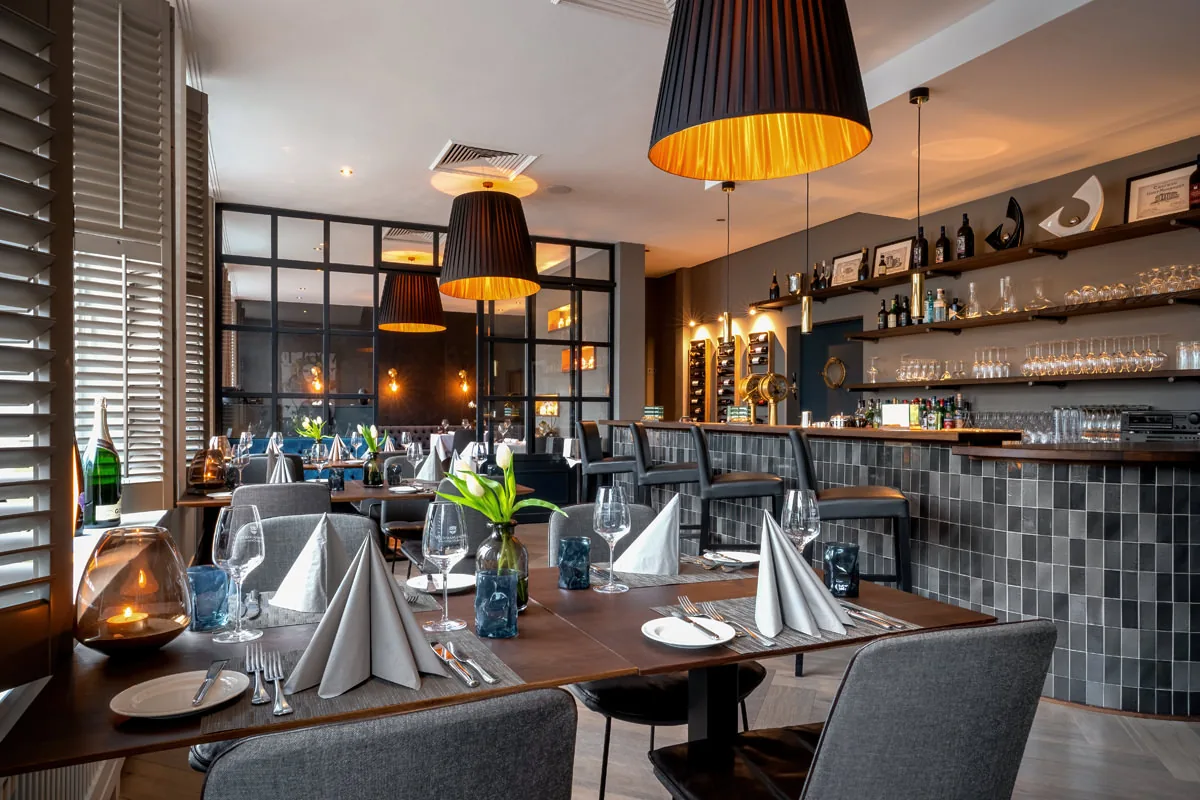 Hotelrestaurant am Abend – Reihe plissierter Leuchten über eingedeckten Tischen, Bar im Hintergrund