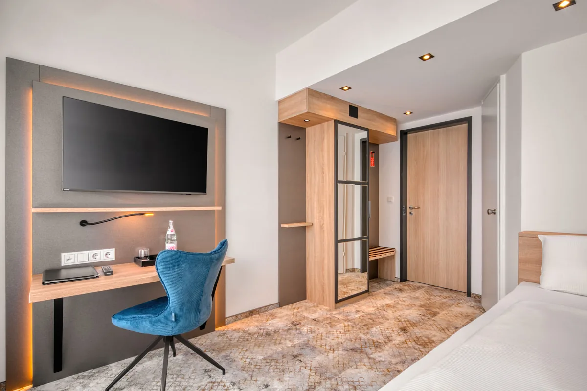 Hotelzimmer mit Wandfernseher, Schreibtisch mit blauem Stuhl und offener Garderobe