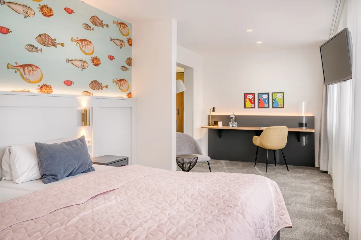 Hotelzimmer mit Fischtapete am Kopfende, langem Schreibtisch und farbigen Grafiken