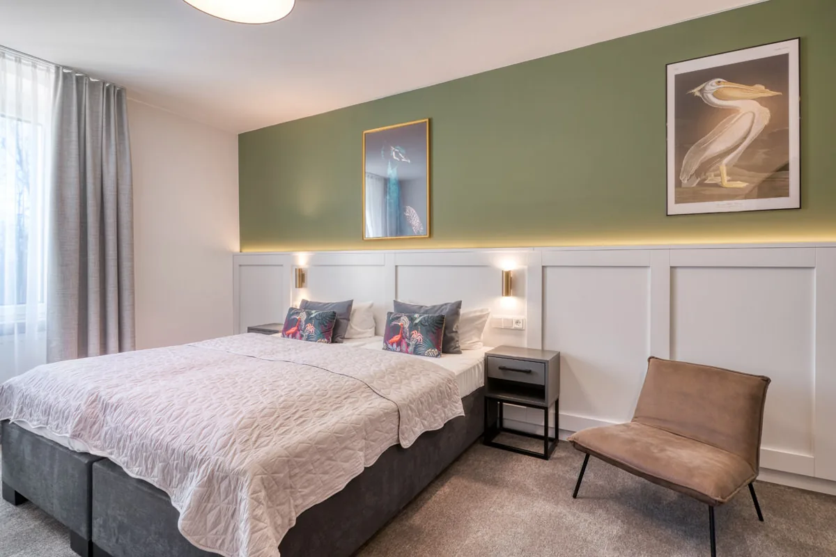 Hotelzimmer mit olivgrüner Wand, gerahmtem Pelikanbild und Boxspringbett mit Tagesdecke