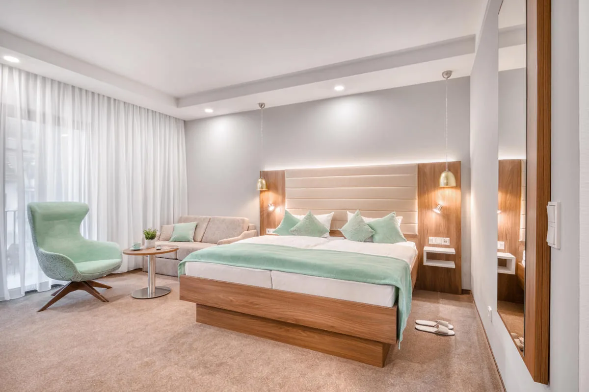 Hotelzimmer in Mint und hellem Holz mit Ohrensessel, Sofa und indirekt beleuchtetem Kopfteil