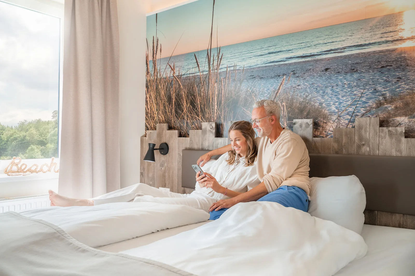 Paar sitzt im Bett vor einem großformatigen Strandmotiv am Kopfende
