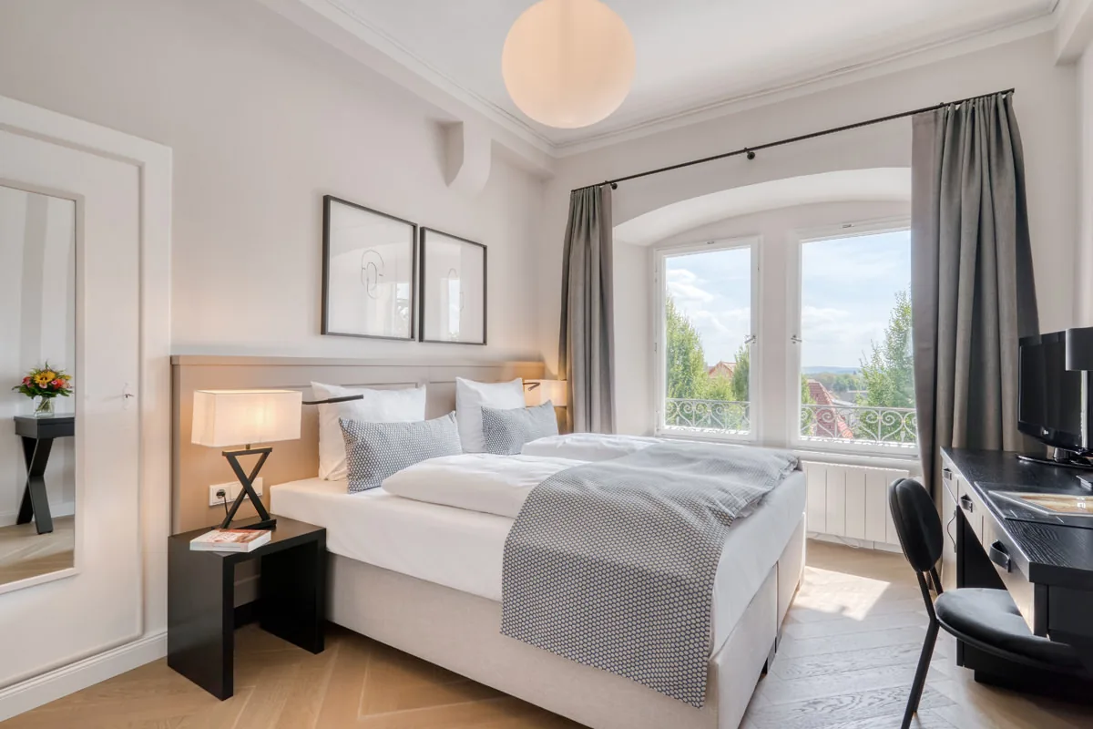 Hotelzimmer mit Rundbogenfenster, Stuckdecke und Kugelleuchte über dem Doppelbett