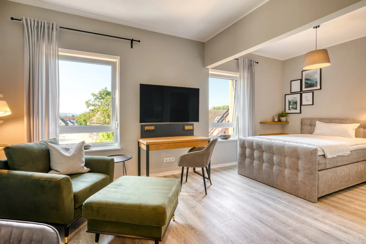 Geräumiges Hotelzimmer mit grüner Sitzecke, Arbeitstisch und Boxspringbett zwischen zwei Fenstern