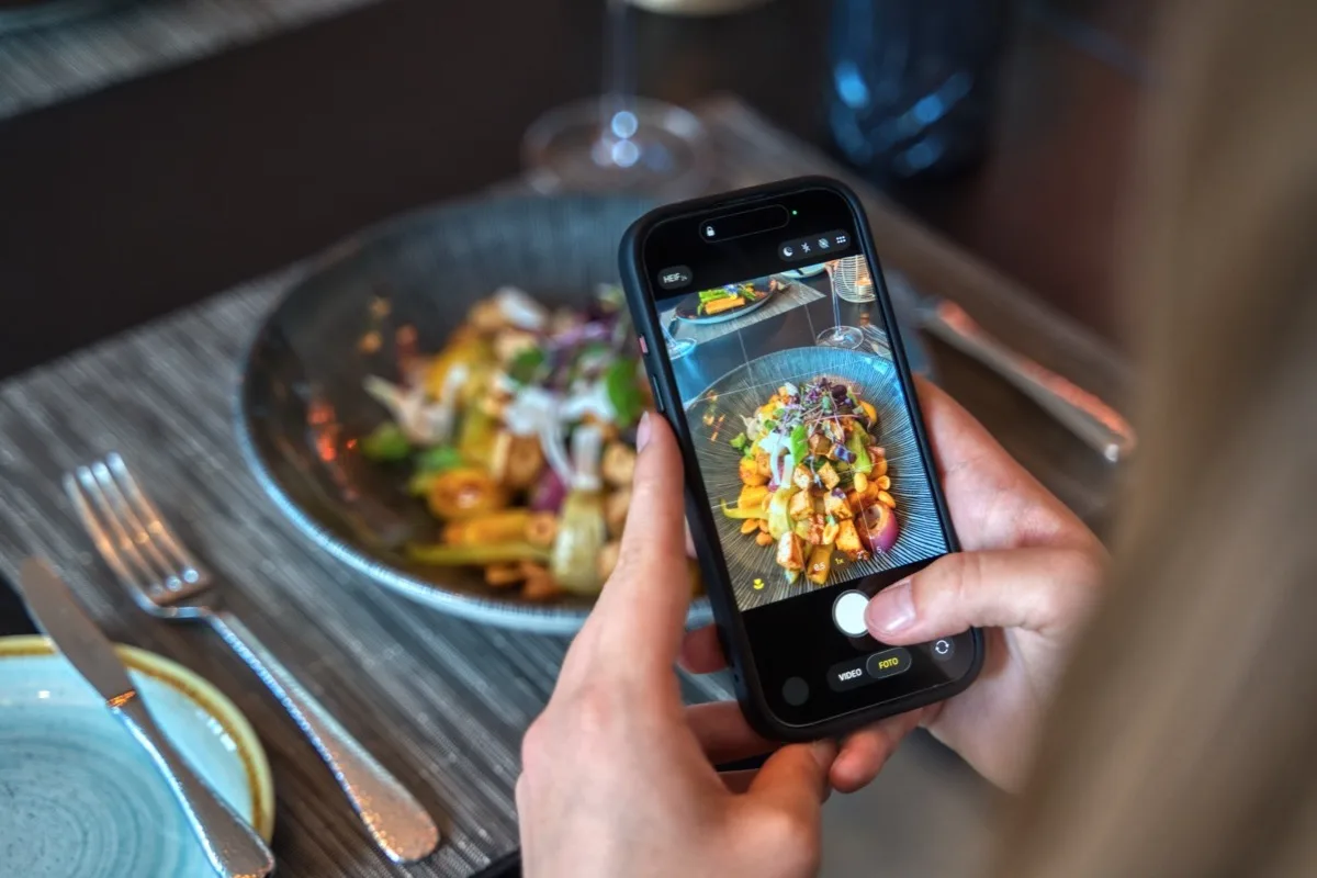 Gast fotografiert seinen Teller mit dem Handy – angerichtetes Gericht in dunkler Schale auf dem Restauranttisch
