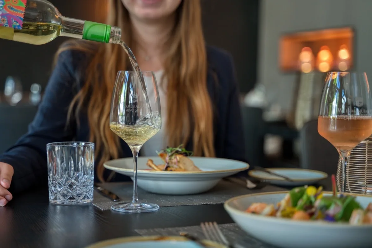 Weißwein wird am gedeckten Tisch eingeschenkt, Gastgeberin im Hintergrund