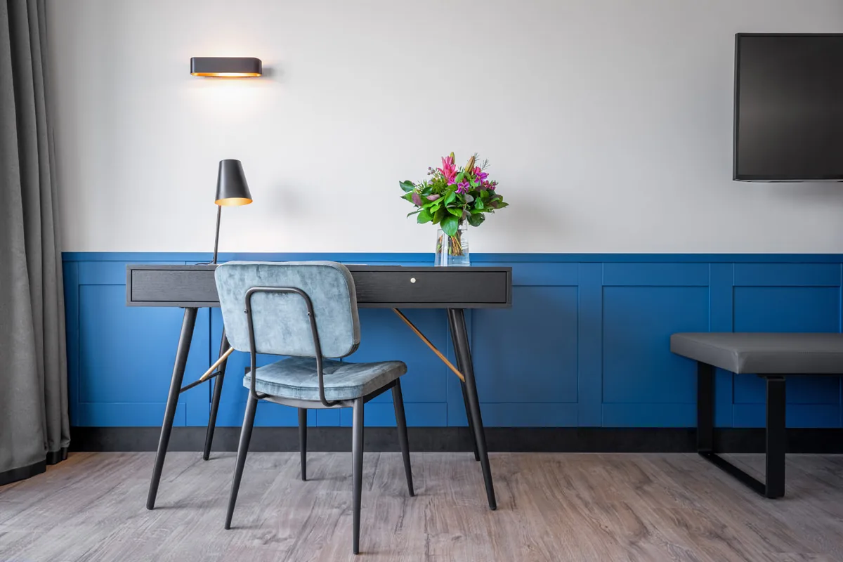 Arbeitsplatz im Hotelzimmer vor blauer Wandvertäfelung mit Leselampe und Blumen